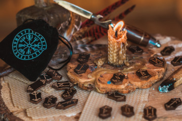 Steine mit leuchtenden, nordischen Runen für Rollenspiel liegen zusammen mit einem schwarzen Beutel und einer Kerze auf einem Holztisch.