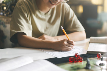 Eine junge Frau sitzt an einem Tisch mit Würfeln und macht Notizen vom Pen and Paper Spiel.